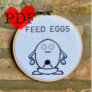 Puede incluir: Un aro de bordado blanco con bordado azul de un personaje de dibujos animados con el texto "FEED EGGS".