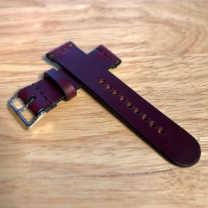 May include: A burgundy leather watch strap with a silver buckle. The strap has multiple holes for adjusting the fit.