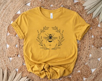 Pode incluir: Uma camiseta amarela mostarda com uma ilustração em preto e branco de uma abelha em uma coroa de folhas. O texto "bee" está impresso abaixo da abelha.