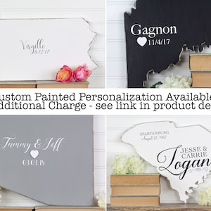 May include: Four custom painted state-shaped wooden signs with distressed edges. The signs are painted in white, gray, black, and light blue. Each sign features a couple's names and a date. The signs are displayed on a shelf with books.