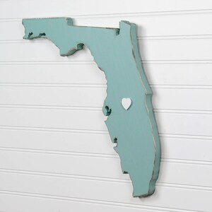 May include: A teal blue wooden cutout of the state of Florida with a white heart placed on the map. The map is mounted on a white wall.