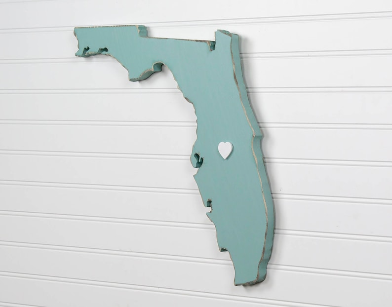 May include: A teal blue wooden cutout of the state of Florida with a white heart placed on the map. The map is mounted on a white wall.