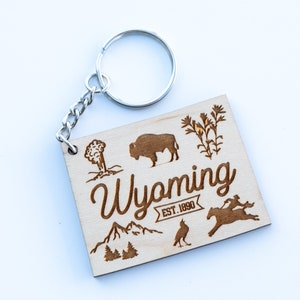 May include: Wooden keychain with a brown silhouette of a bison, a geyser, a mountain range, a bird, and a horse. The text "Wyoming EST. 1890" is engraved on the keychain.