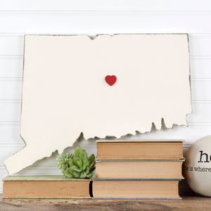 May include: A white wooden outline of the state of Connecticut with a red heart in the center. The outline is sitting on a stack of books and a white round object with the word "home" on it. The text "This is where your journey begins" is on the round object.