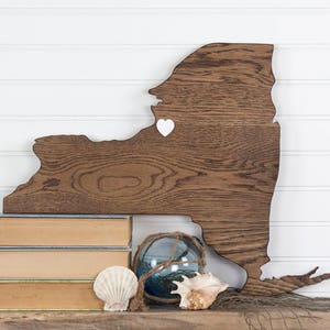 May include: A wooden cutout of the state of New York with a white heart in the center. The cutout is sitting on a stack of three books and a decorative glass orb. There are also two seashells in the image.