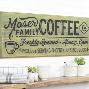 May include: A green wooden sign with black text that reads "Moser Family Coffee Co. Freshly Brewed - Always Open Proudly Serving Phoenix, AZ Since 2006". The sign has a stylized coffee cup with steam coming from it.