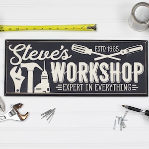 May include: Black and white sign with the text "Steve's Workshop - Expert in Everything" and the year "ESTD 1965". The sign features a hammer, screwdriver, and drill.