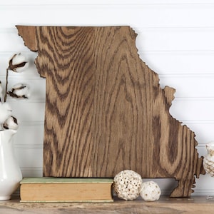 May include: A wooden cutout of the state of Missouri, stained in a dark brown color. The cutout is displayed on a white shelf with a book, a vase of cotton bolls, and decorative white orbs.