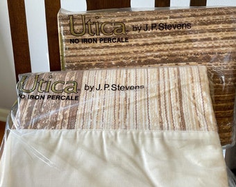 May include: Two new, packaged Utica brand twin flat sheets. The top portion of the sheets features a brown and beige striped pattern, while the bottom is a solid cream color. The packaging reads "NO IRON PERCALE".