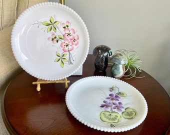 May include: Two white ceramic plates with pearl-like edges and hand-painted floral designs. The plates feature a pink and green floral design and a purple and green floral design.