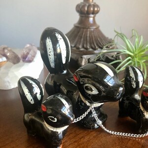 May include: Three black and white ceramic skunk figurines. The skunks are in a line, with the middle skunk holding a silver chain. The skunks have white stripes and red noses.