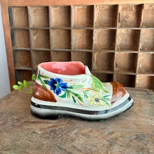 May include: A ceramic shoe-shaped planter with a brown toe and heel, white body, and black sole. The shoe is decorated with blue, yellow, and green floral designs. The interior is pink. The planter sits on a wooden surface.
