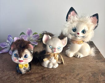 May include: Three vintage ceramic cat figurines with fluffy fur. The largest cat is white with black ears and a gold collar. The middle cat is white with black ears and a yellow bow. The smallest cat is brown with black ears and a yellow bow.
