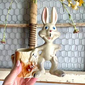 May include: A vintage ceramic planter shaped like a rabbit with a tree stump base. The rabbit is white with blue eyes and a pink nose. The tree stump is brown and has a light brown top.