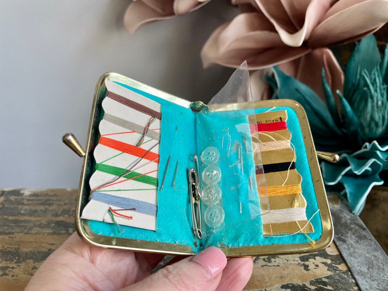 Puede incluir: Un peque&ntilde;o kit de costura de metal dorado con un forro turquesa. El kit est&aacute; abierto y contiene carretes de hilo, agujas y botones. El texto "MADE IN" es visible en el lado derecho del kit.