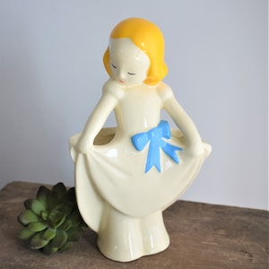 May include: A vintage ceramic planter shaped like a girl with yellow hair, wearing a white dress with a blue bow. The planter is holding her dress out, creating a space for a plant.