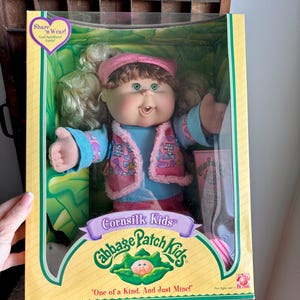 May include: A Cabbage Patch Kids Cornsilk Kids doll in original packaging. The doll has brown hair, green eyes, and wears a blue shirt with a pink patterned vest and matching pink visor. The box features the text Share 'n Wear and One of a Kind.