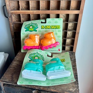 May include: Two sets of Cabbage Patch Kids toy skates in original packaging. The top set features orange boots with bright pink wheels. The bottom set features teal boots with white blades. Both packages are light green with the Cabbage Patch Kids logo.