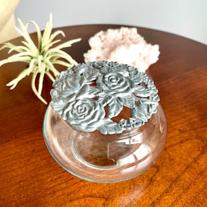 May include: A clear glass jar with a silver lid featuring a floral design. The lid has a raised design of roses and leaves.