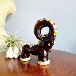 May include: A brown ceramic ram figurine with painted details. The ram has a large, curled horn with colorful stripes. The figurine is standing on a wooden surface.