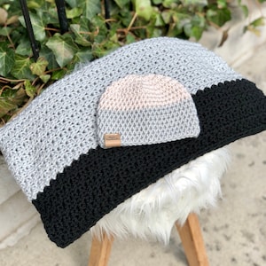 Op de afbeelding: Een grijs en zwart gehaakt deken met een bijpassende grijs en roze gehaakte muts. De muts heeft een klein bruin label met het woord "Handmade" erop.