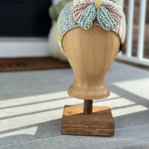 Puede incluir: Una diadema de crochet con un gran lazo en tonos verdes, amarillos y rosas. La diadema se muestra en un maniquí de madera.