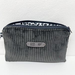 Puede incluir: Un estuche de pana gris con cierre de cremallera y un pequeño lazo para llavero. El forro interior presenta un estampado de animales en blanco y negro. Una pequeña etiqueta de cuero en la parte delantera dice "HAND MADE".