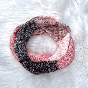 May include: A knitted infinity scarf in shades of pink, brown, and black. The circular scarf is made of a soft, textured yarn and is displayed on a white, fluffy surface. The color-blocked design adds visual interest to this cozy accessory.
