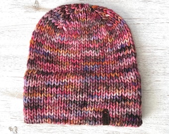 May include: A chunky knit beanie featuring a variegated yarn pattern in shades of pink, purple, orange, and dark brown. The hat has a folded brim and a small rectangular brown leather tag attached to the lower right side with the text Alaskan Knits.