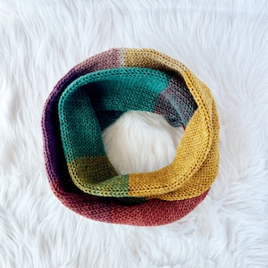 May include: A knitted infinity scarf in a circular shape, featuring a color block design. The scarf showcases a gradient of colors, including shades of purple, teal, gold, and red. The soft texture and warm tones suggest a cozy accessory.