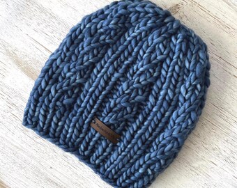 May include: A chunky knit beanie in a deep blue color, featuring a prominent cable knit pattern throughout the fabric. A small, rectangular brown leather tag is attached to the lower front, embossed with the text AlaskanKnitCo.