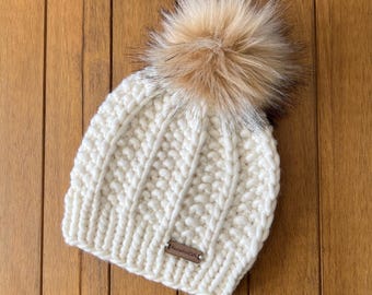 May include: A chunky knit beanie in off-white yarn featuring a ribbed texture and a large, fluffy faux fur pom-pom in shades of tan and brown. A small, rectangular brown leather-like tag is attached to the lower edge of the knit hat.