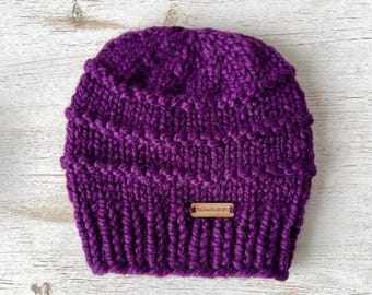 May include: A deep purple chunky knit beanie hat featuring a ribbed brim and a textured stitch pattern. A small rectangular wooden tag is attached to the front, clearly engraved with the text AlaskanKnitGifts in a dark brown font.