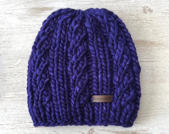 May include: A deep purple beanie crafted from thick, chunky yarn featuring a prominent vertical cable knit pattern. A small, rectangular brown leather tag is stitched onto the lower right side, clearly displaying the text AlaskanKnitGifts.