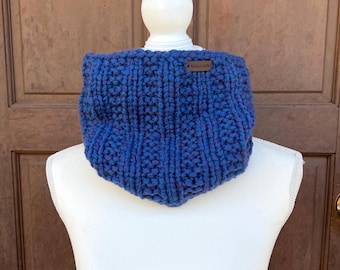 May include: A chunky knit cowl in a deep blue yarn with a ribbed texture. The cowl features a small rectangular brown leather tag attached to the upper right side with the text "Handmade with Love" clearly visible on the surface.