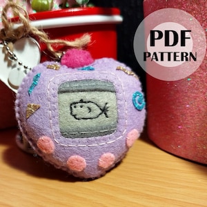 May include: Purple heart-shaped plush toy with a screen that says "PDF PATTERN" and a small fish drawn on the screen. The toy is a felt version of a Tamagotchi.