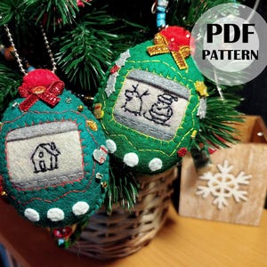 May include: Two handmade Christmas ornaments with felt details. One is teal with a house design, the other is green with a reindeer and snowman design. Both have red bows and are hanging on a Christmas tree.