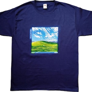 Könnte beinhalten: Marineblaues T-Shirt mit einem farbenfrohen Aufdruck, der eine grüne Wiese, einen blauen Himmel und weiße Wolken zeigt, die an den Windows XP-Desktop-Hintergrund erinnern. Der Text "Windows XP 2001" ist sichtbar. Ein klassisches T-Shirt.