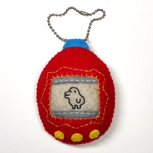 K&ouml;nnte beinhalten: Roter Filz-Schl&uuml;sselanh&auml;nger in Form eines Tamagotchis mit blauer Oberseite und silberner Kette. Der Bildschirm zeigt ein kleines wei&szlig;es Wesen mit schwarzen Augen.