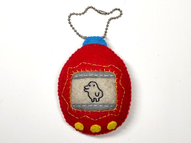 K&ouml;nnte beinhalten: Roter Filz-Schl&uuml;sselanh&auml;nger in Form eines Tamagotchis mit blauer Oberseite und silberner Kette. Der Bildschirm zeigt ein kleines wei&szlig;es Wesen mit schwarzen Augen.