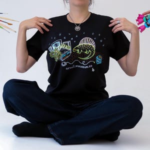 Könnte beinhalten: Schwarzes T-Shirt mit einer farbenfrohen Grafik mit einem Dinosaurier, einem Computer und Text. Das T-Shirt wird von einer Person getragen, die im Schneidersitz sitzt. Pinsel und Süßigkeiten werden auf beiden Seiten gehalten.