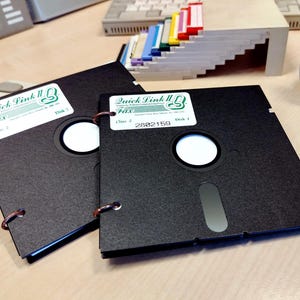 May include: Two black 3.5-inch floppy disks with labels reading "Quick Link II Fax". The disks are bound together with copper rings. One label reads "Disk 1" and the other reads "Disk 2". The disks are on a wooden desk.
