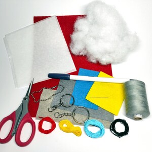 K&ouml;nnte beinhalten: Ein Bastelset zum Herstellen eines Schl&uuml;sselanh&auml;ngers. Das Set enth&auml;lt Filz in Rot, Blau, Gelb und Grau, einen wei&szlig;en Stift, eine Nadel, Garn, eine Schere, einen Schl&uuml;sselring und F&uuml;llmaterial.