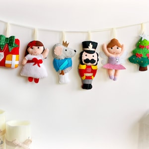 May include: A felt Christmas garland featuring a red gift box, a girl in a white dress, a mouse in a blue coat, a nutcracker, a ballerina, and a green Christmas tree.