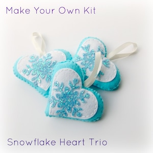 Può includere: Tre ornamenti a forma di cuore in feltro blu con centri in feltro bianco e disegni di fiocchi di neve blu scintillanti. Gli ornamenti hanno anelli di nastro bianco per appendere. Il testo "Make Your Own Kit" e "Snowflake Heart Trio" è visibile nell'immagine.