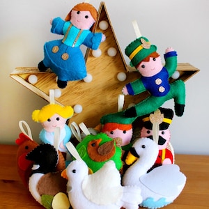 May include: A set of felt ornaments featuring a variety of characters, including a girl in a blue dress, a boy in a green suit, a bird, a swan, and a rooster. The ornaments are hanging from a wooden star.
