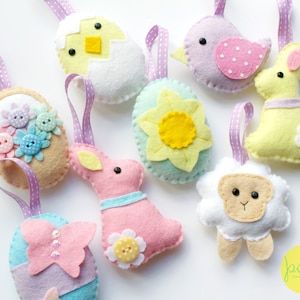 Könnte beinhalten: Neun Filz-Ostereier mit Aufhängeschlaufen. Die Anhänger zeigen ein Küken im Ei, einen Vogel, ein Häschen, ein Lamm, eine Blume und drei Eier mit verschiedenen Designs.