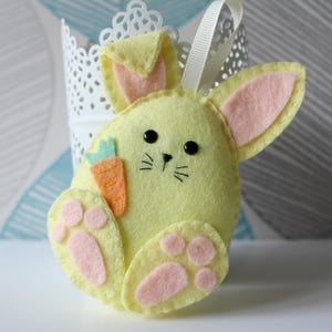 May include: A yellow felt Easter bunny decoration with pink inner ears and paw pads. The bunny has black eyes, whiskers, and a small carrot. A white ribbon is attached for hanging. A charming Easter-themed ornament.