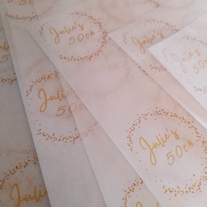 May include: A stack of translucent white paper sheets with gold lettering that reads "Julie's 50th" in a circle with gold glitter.