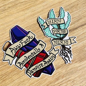 May include: Two stickers on a wooden surface. One sticker features a purple and red potion bottle with the text "Aww Fuck Accidentally A Vampire Again". The other sticker has a plant with the text "YIRN FOR THE NIRN".